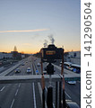 Camera On Tripod Overlooking Busy Highway At Sunset Capturing Dusk Traffic And Urban Commute 141290504