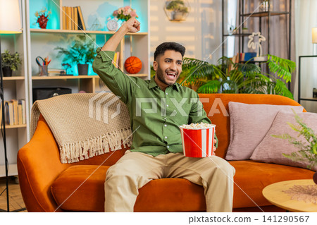 Indian man on home sofa cheering football goal with popcorn while watching match and celebrating win Indian man on home sofa cheering football goal with popcorn while watching match and celebrating win 141290567