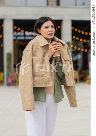 Young woman feeling cold walking in city street wrapped in jacket while sipping hot drink outdoors Young woman feeling cold walking in city street wrapped in jacket while sipping hot drink outdoors 141290647