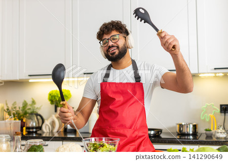 Arabian young adult man dancing in kitchen mixing fresh salad while listening to music in headphones Arabian young adult man dancing in kitchen mixing fresh salad while listening to music in headphones 141290649