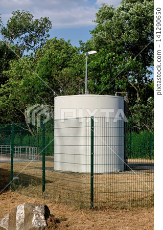 White industrial storage tank enclosed by green mesh fence with trees in background White industrial storage tank enclosed by green mesh fence with trees in background 141290650