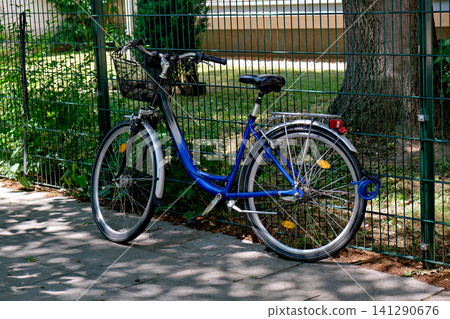 Blue city bicycle with basket parked next to green fence Blue city bicycle with basket parked next to green fence 141290676
