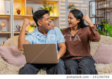 Indian couple man and woman sitting on home sofa celebrating approval on laptop with joyful victory Indian couple man and woman sitting on home sofa celebrating approval on laptop with joyful victory 141290769