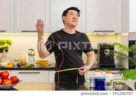 Heavy Asian adult man measures waist with tape upset about big belly fat weight gain at home kitchen 141290797