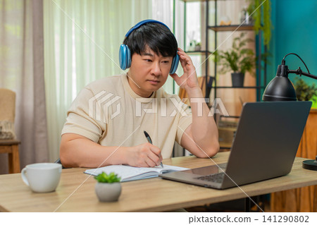 Asian man listening to podcast on laptop headphones, nodding and writing detailed notes in notebook Asian man listening to podcast on laptop headphones, nodding and writing detailed notes in notebook 141290802