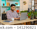 Indian man on home table working on laptop drinking coffee smiling during focused remote work task 141290821