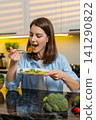 Young woman eating healthy salad in kitchen smiling with fork during nutrition lifestyle meal break 141290822