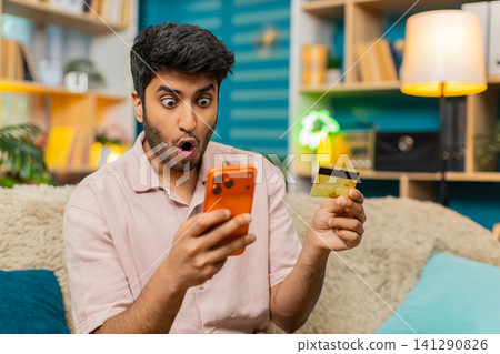 Indian man on home sofa upset after payment decline checking bank card and smartphone in frustration Indian man on home sofa upset after payment decline checking bank card and smartphone in frustration 141290826