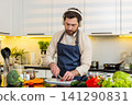 Middle-aged man singing with headphones while slicing red pepper for healthy salad in home kitchen 141290831