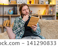 Young man on home sofa reading book with happy smile and enjoying peaceful leisure time in comfort 141290833