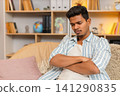 Indian man on home sofa frowning with dissatisfied look while sitting in thought in cozy living room 141290835