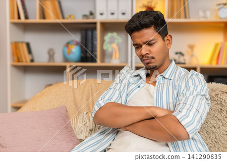 Indian man on home sofa frowning with dissatisfied look while sitting in thought in cozy living room Indian man on home sofa frowning with dissatisfied look while sitting in thought in cozy living room 141290835