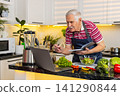 Senior old man making vegetable salad using laptop recipe and writing notebook notes for diet plan 141290844