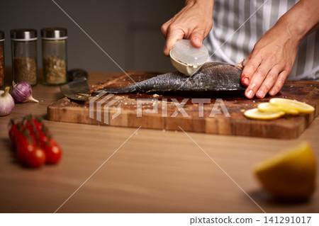 Person wearing an apron scaling fresh dorado fish with a scaler on a wooden cutting board in a kitchen Person wearing an apron scaling fresh dorado fish with a scaler on a wooden cutting board in a kitchen 141291017