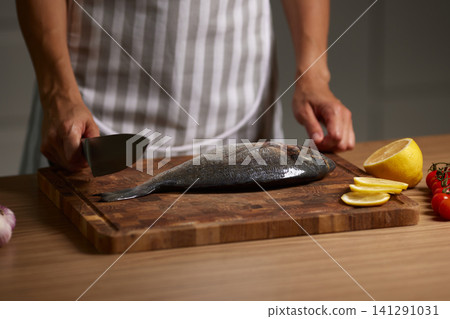 Chef's hands holding a knife, preparing a whole fresh sea bream on a wooden cutting board with lemon and tomatoes Chef's hands holding a knife, preparing a whole fresh sea bream on a wooden cutting board with lemon and tomatoes 141291031