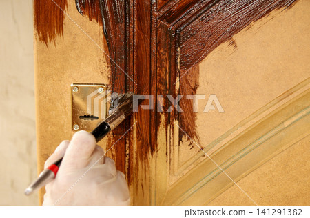 Close up of hand in glove painting wooden door with brown varnish or stain. Person restoring old wood door with paintbrush, home renovation, DIY improvement and woodwork concept Close up of hand in glove painting wooden door with brown varnish or stain. Person restoring old wood door with paintbrush, home renovation, DIY improvement and woodwork concept 141291382