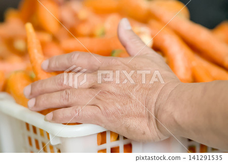 Closeup of a persons hand resting on a plastic crate filled with fresh, raw carrots in a market or farm setting Closeup of a persons hand resting on a plastic crate filled with fresh, raw carrots in a market or farm setting 141291535