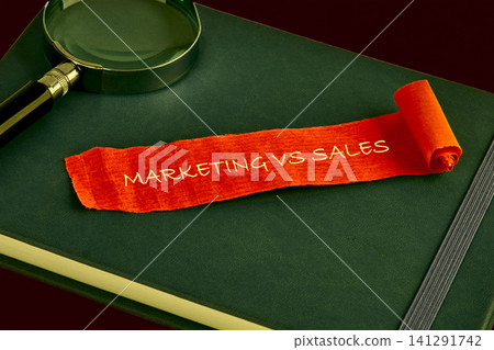 Bright orange paper scroll featuring the words Marketing vs Sales sits on a dark green notebook next to a magnifying glass symbolizing an investigative look into commercial growth Bright orange paper scroll featuring the words Marketing vs Sales sits on a dark green notebook next to a magnifying glass symbolizing an investigative look into commercial growth 141291742