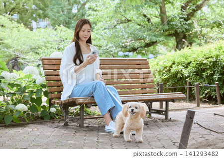 一位年輕女子帶著她的狗坐在公園長椅上,看著她的智慧型手機。 一位年輕女子帶著她的狗坐在公園長椅上,看著她的智慧型手機。 141293482