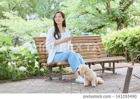 一位年輕女子帶著她的狗坐在公園長椅上，看著她的智慧型手機。 141293490
