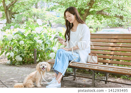 一位年輕女子帶著她的狗坐在公園長椅上,看著她的智慧型手機。 一位年輕女子帶著她的狗坐在公園長椅上,看著她的智慧型手機。 141293495