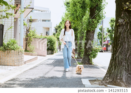 A young woman with a smile walks her dog in a residential area surrounded by fresh greenery. A young woman with a smile walks her dog in a residential area surrounded by fresh greenery. 141293515