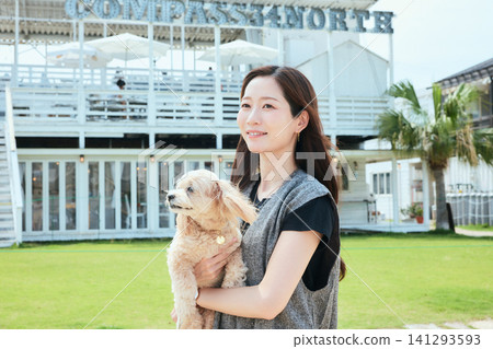 一位面帶微笑的女士抱著她的狗站在海邊咖啡館的草坪上。 一位面帶微笑的女士抱著她的狗站在海邊咖啡館的草坪上。 141293593