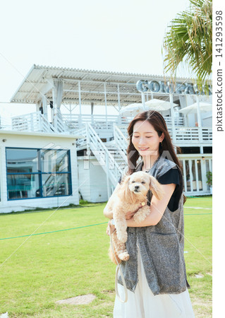 A young woman smiling while holding her dog, in a stylish cafe with a lush green lawn. A young woman smiling while holding her dog, in a stylish cafe with a lush green lawn. 141293598