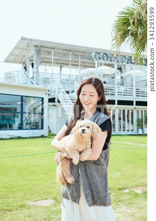 A young woman smiles, holding her dog, in front of a dog-friendly cafe with a lush green lawn. A young woman smiles, holding her dog, in front of a dog-friendly cafe with a lush green lawn. 141293599