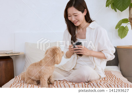A smiling young woman relaxing on a bed with her dog, holding a mug. A smiling young woman relaxing on a bed with her dog, holding a mug. 141293627