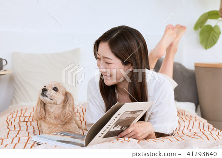 A young woman with a smile is lying in bed, reading a magazine with her dog. A young woman with a smile is lying in bed, reading a magazine with her dog. 141293640