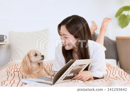 一位微笑的年輕女子躺在床上,和她的狗一起看雜誌。 一位微笑的年輕女子躺在床上,和她的狗一起看雜誌。 141293642