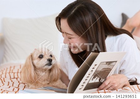 A young woman smiles as she reads a magazine with her dog on the bed. 141293651