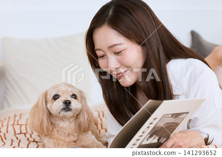 一位年輕女子帶著微笑躺在床上,一邊看雜誌一邊和她的狗狗玩耍。 一位年輕女子帶著微笑躺在床上,一邊看雜誌一邊和她的狗狗玩耍。 141293652