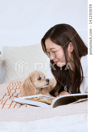 A young woman with glasses reading a magazine on the sofa with her dog. 141293659