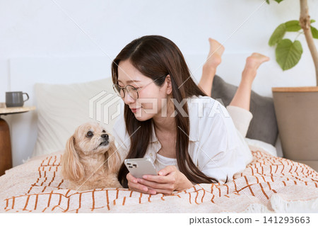 A woman wearing glasses is lying in bed, looking at her smartphone while spending time with her dog. A woman wearing glasses is lying in bed, looking at her smartphone while spending time with her dog. 141293663