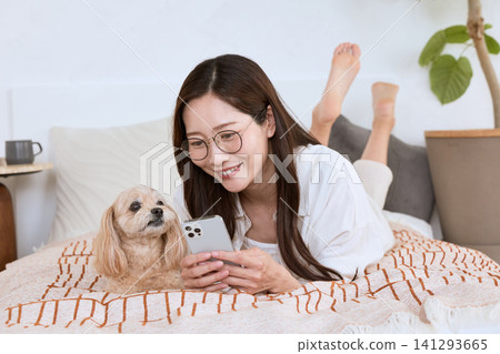 A woman wearing glasses is lying on a bed looking at her smartphone, and her dog is also there. A woman wearing glasses is lying on a bed looking at her smartphone, and her dog is also there. 141293665