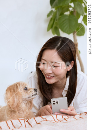 A young woman smiles as she relaxes on the sofa with her dog, looking at her smartphone. 141293701