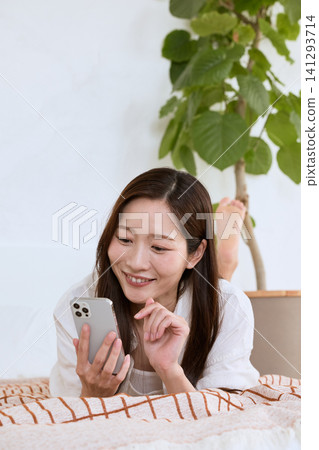 A young woman smiling while looking at her smartphone in the living room. A young woman smiling while looking at her smartphone in the living room. 141293714