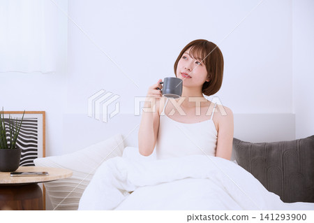 A young woman relaxing on a bed, drinking coffee. 141293900