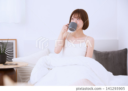 清晨,一位年輕女子躺在床上,手捧咖啡杯,正在喝咖啡。 清晨,一位年輕女子躺在床上,手捧咖啡杯,正在喝咖啡。 141293905