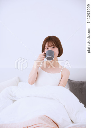 清晨,一位年輕女子躺在床上,手裡拿著杯子喝咖啡。 清晨,一位年輕女子躺在床上,手裡拿著杯子喝咖啡。 141293909