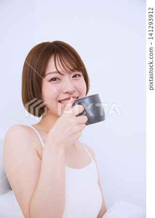 一位身穿白色洋裝的年輕女子手捧一杯飲品，面帶微笑，悠閒地享受著時光。 141293912