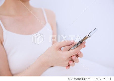 一位身穿白色吊帶背心的年輕女子正在操作她的智慧型手機。 141293921