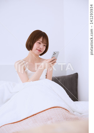 一位年輕女子坐在床上使用智慧型手機。 141293934