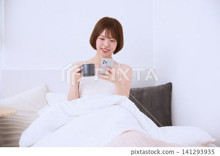 一位面帶微笑的年輕女子躺在床上,手裡拿著智慧型手機和一杯咖啡。 一位面帶微笑的年輕女子躺在床上,手裡拿著智慧型手機和一杯咖啡。 141293935