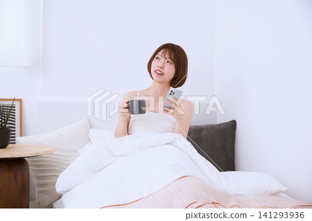 年輕女性的早晨日常:躺在床上,一邊玩手機一邊喝咖啡。 年輕女性的早晨日常:躺在床上,一邊玩手機一邊喝咖啡。 141293936