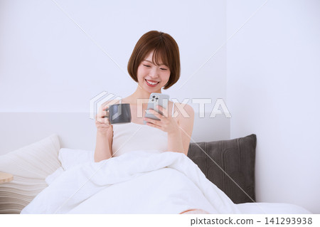 一位年輕女子麵帶微笑,躺在床上一邊喝咖啡一邊看著她的智慧型手機。 一位年輕女子麵帶微笑,躺在床上一邊喝咖啡一邊看著她的智慧型手機。 141293938