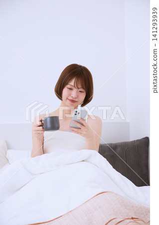 一位年輕女子坐在床上,手裡拿著智慧型手機和馬克杯。 一位年輕女子坐在床上,手裡拿著智慧型手機和馬克杯。 141293939
