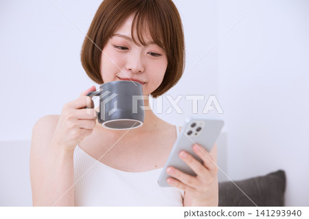 一位年輕女子一邊喝咖啡一邊看著她的智慧型手機。 一位年輕女子一邊喝咖啡一邊看著她的智慧型手機。 141293940